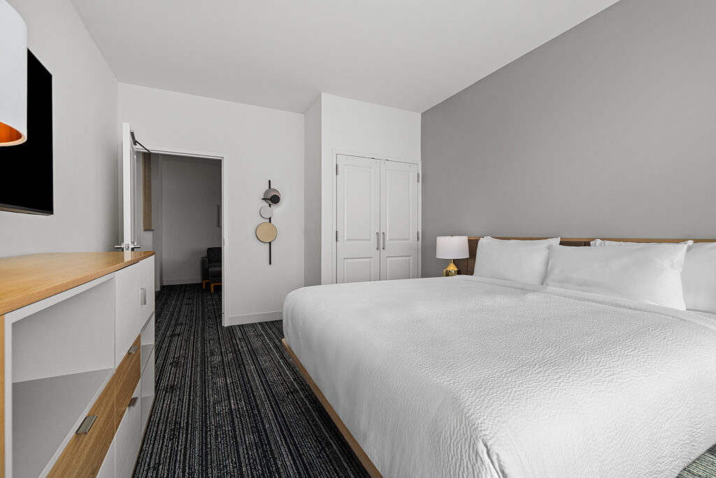 Modern TownePlace Suites hotel room in TN featuring a large neatly made bed with white linens, bedside lamp, double-door closet, dresser, and a view into a sitting area. The minimal decor showcases neutral colors for a relaxing stay.