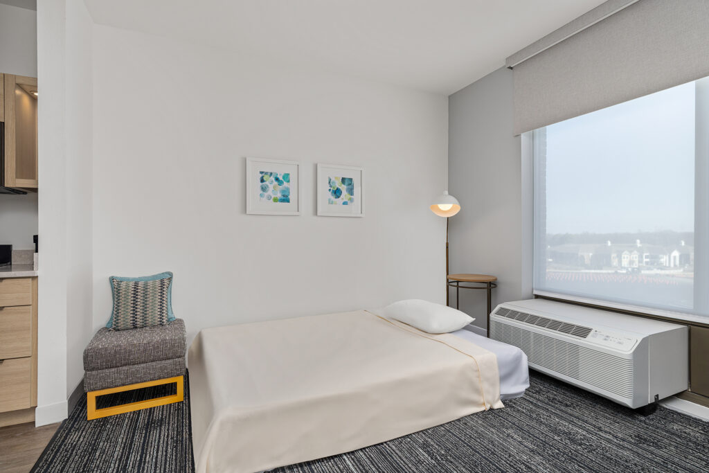 A minimalist bedroom inspired by hotel hospitality, featuring a low bed with light beige bedding, modern lamp, abstract framed artworks on a white wall, a gray chair with a colorful cushion, and a large window offering a scenic view outside.
