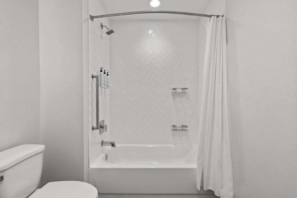 A modern white bathroom at TownePlace Suites in Murfreesboro features a bathtub with a white tile herringbone pattern, shower curtain, overhead shower, wall-mounted toiletries, and metal shelves. A toilet is visible on the left—hotel comfort at its best.