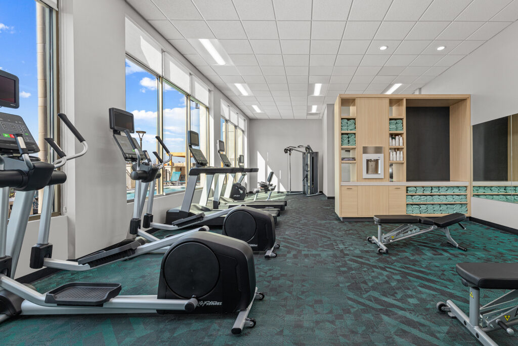 A modern gym in the hotel with treadmills, elliptical machines, benches, free weights, mirrors, towel shelves, and large windows letting in natural light—offering guests top-tier hospitality and fitness amenities.