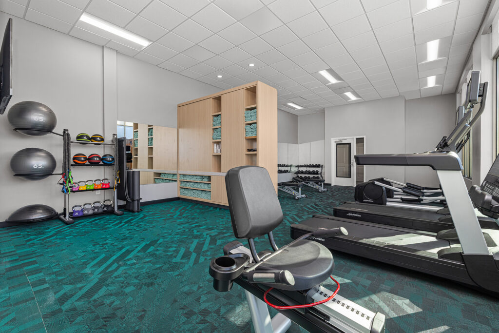 A modern hotel gym in Murfreesboro, TN with a stationary bike, treadmill, free weights, medicine balls, exercise mats, and mirrored walls. Bright lighting, teal-patterned carpet, and wooden storage shelves with towels complete the space.