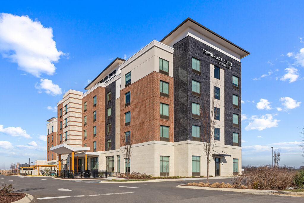 A modern, multi-story hotel building labeled TownePlace Suites, showcasing hospitality with brick, cream, and dark gray exterior walls, large windows, and a covered entrance under a bright blue sky with scattered clouds.